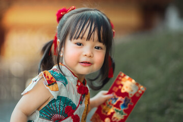 Close-up Happy Asian toddler girl in traditional Chinese dress Qipao smiling and holding a red envelope (Angpao). Chinese New Year celebration childhood joy and cultural tradition concept.