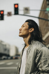 Calm East Asian man in light grey bomber jacket practicing mindful commuting while waiting at a crosswalk against a blurred grey urban background.
