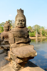 Obraz premium Close-up of a weathered stone guardian figure on the monumental bridge leading to Angkor Thom near Siem Reap, Cambodia. The expressive sculpture stands beside a tranquil moat, framed by tropical