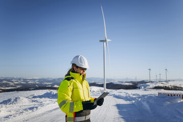 Female engineer standing in large field energy windmills using Digital Tablet and inspecting wind turbines. Wind turbine in background. Wind power generation.