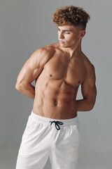 Obraz premium Young muscular man standing shirtless with white pants on gray background showing athletic body and defined muscles with curly hair and pleasant facial expression