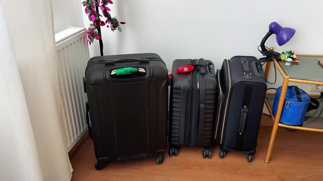 Three suitcases - one large and two medium - stand lined up in a bright room next to a side table. A blue cooler bag sits below a purple lamp, capturing the quiet anticipation of travel