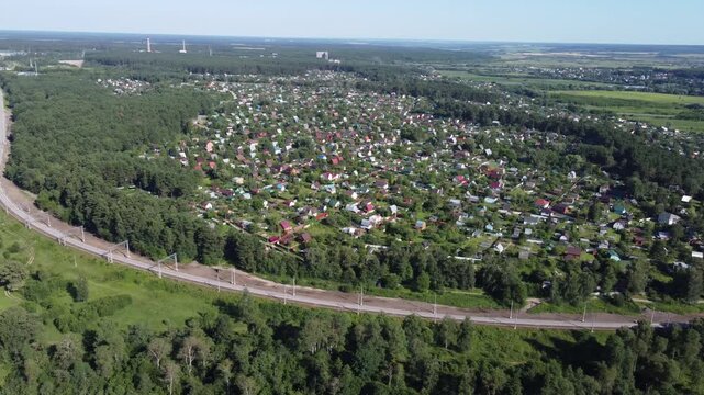 Aerial panorama of a vibrant dacha community and active railway snaking through verdant forests, with distant fields under a bright summer sky