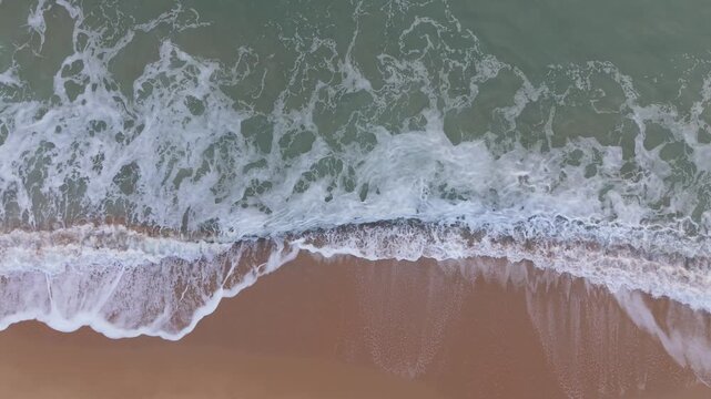 Top down drone view of winter surf rolling onto empty beach with chilly water tones forming calm atmospheric seascape