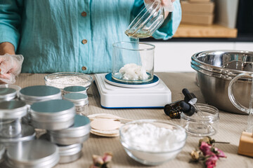 Hand in glove pours liquid oil from bowl into beaker containing white solid butter on digital scale. cosmetic formulation process, oil phase emulsion, cream texture transition, handmade moisturizer.