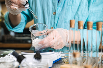 Person in gloves uses glass pipette to dispense liquid into beaker with white powder on digital scale. lab-style skincare, botanical chemistry aesthetic, diy cosmetic formulation, apothecary jars.