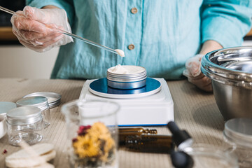 Person in gloves uses glass rod to apply cream into metal tin on digital scale. Botanical ingredients aesthetic, raw beauty materials, herbal mixology, natural skincare components.