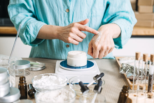 Person applies white cream to back of hand above jar on digital scale surrounded by skincare ingredients. tactile beauty DIY, silk-feel cream, sensory skincare testing, textural wellness ritual.