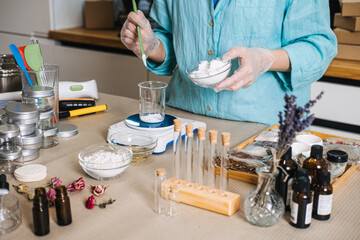 Person adds white powder from bowl into glass beaker on electronic scale surrounded by botanical ingredients. Lab-style skincare, botanical chemistry, diy cosmetic formulation, apothecary jars.