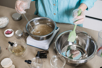 Person uses glass rod to stir liquid in metal bowl on digital scale while holding green spatula. Precise cosmetic formulation process, aesthetic beauty lab scale, artisan skincare manufacturing.