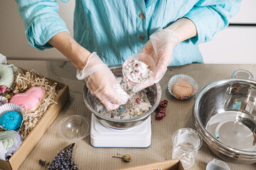 Hands mix white powder and dried botanicals in metal bowl on digital scale surrounded by finished products. Handmade bath bombs process, cosmetic chemistry at home, artisan soap making.