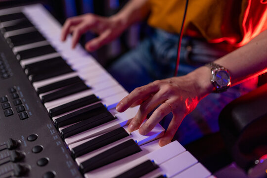Close up of hands playing MIDI keyboard. Musician composing music on piano keys in dark studio with neon blue and red lights. Creative audio production and electronic music concept background.