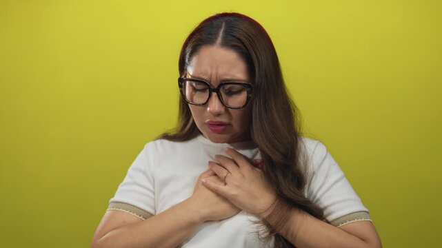 Woman clutch chest wearing tortoiseshell glasses and white tee with long hair in studio for heartattack; concern.