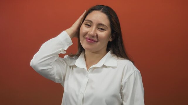 Young hispanic woman in white shirt holds hair in studio with deep red walls; confidence selfcare empowerment grace.