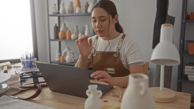 Young hispanic artisan woman wearing brown apron with hand on cheek typing on laptop in studio; thinking creativity focus.