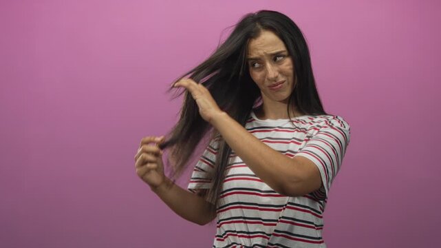 Woman holding and examining long hair for split ends in studio with pink backdrop; haircare frustration.