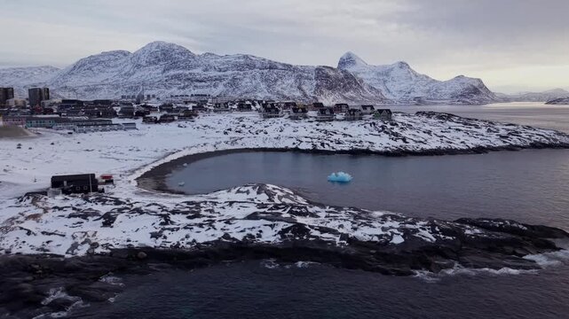 Drone shot of Nuuk, Greenland, featuring colorful houses on the snowy coast, a bright blue iceberg in the calm bay, and majestic Arctic mountains in the background during the winter season.