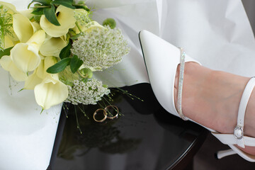 Bride resting her legs in elegant white heels near a window, with a wedding bouquet on a black table. Soft natural light, calm morning atmosphere, bridal preparation concept