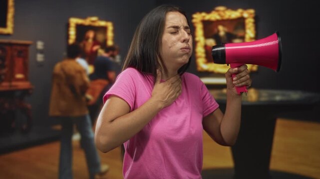 Young hispanic woman shouting into pink megaphone in an art gallery building while gesturing with hand to throat; urgent protest.