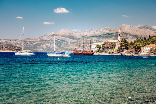 View of azure waters shimmer under the sun, boats bob gently near the shore, while a quaint village nestles against a backdrop of rugged mountains, Lopud, Dubrovnik-Neretva County, Croatia.