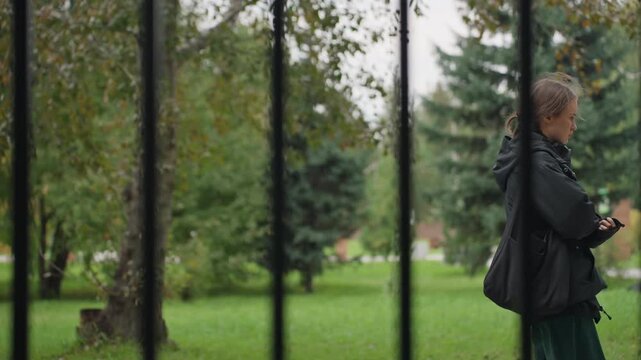 Pensive woman waiting at park fence. Overcast autumn afternoon, woman wearing dark jacket and carrying bag, walking along pathway, pausing to fold arms and look away, candid profile seen through black