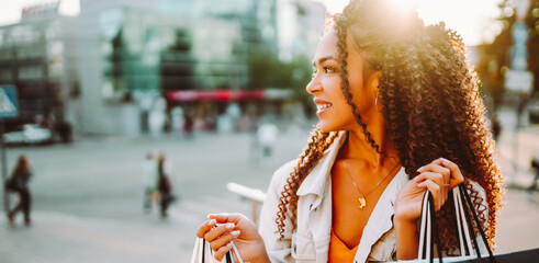 Portrait of a beautiful woman with shopping bags on a city street in the sun's rays. A young woman with a phone enjoys the sunset after shopping. Concepts of consumerism, shopping, and discounts. © maxbelchenko