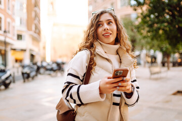 Obraz premium Portrait of a stylish woman with a phone in a busy city square. A beautiful woman enjoys a stroll on a sunny day and blogs on her phone. Lifestyle. Tourism concept.