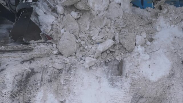 A close-up of a front-end loader bucket collecting ice chunks and frozen snow from the asphalt. Mechanized cleaning of icy road sections to ensure traffic safety