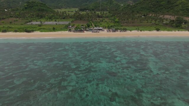 Smooth aerial flight along the coastline of Pantai Lancing and Tampah Beach in southern Lombok, Indonesia. Turquoise water, sandy beach, gentle waves and lush green hills under cloudy tropical skies.