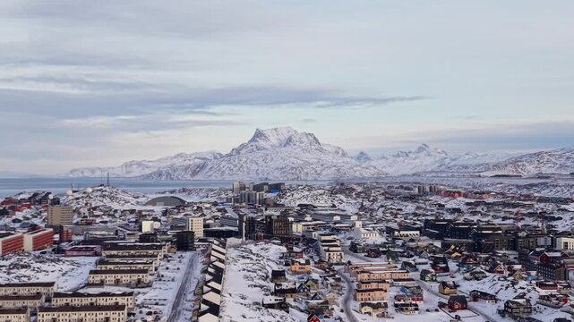 Scenic aerial panorama of Nuuk, Greenland, featuring colorful Nordic houses in a snow-covered landscape, modern city architecture, and the Arctic ocean under a soft winter sunset sky.