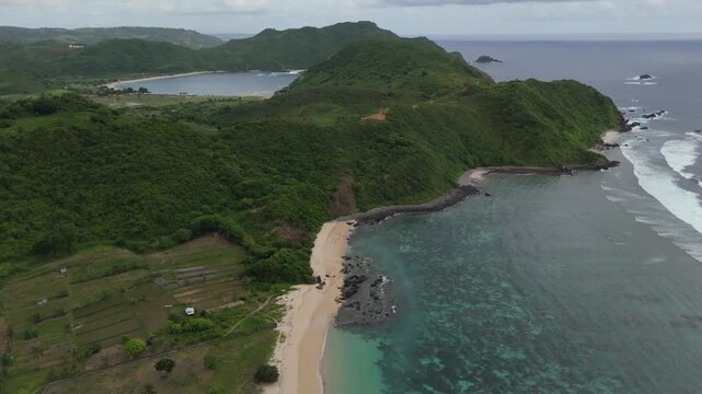 Smooth drone orbit along Pantai Lancing and Tampah Beach in southern Lombok, Indonesia. Turquoise water, sandy shoreline, rolling waves, green hills and local warungs during cloudy rainy season.