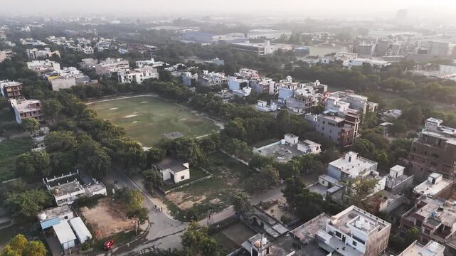 Aerial view of a residential sector in Greater Noida featuring low-rise homes, a central playground, tree-lined streets, and light haze, reflecting calm urban life amid growing city development.