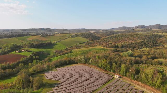 Aerial boom-down revealing large solar panel farm integrated into lush green Tuscan countryside with vineyards.