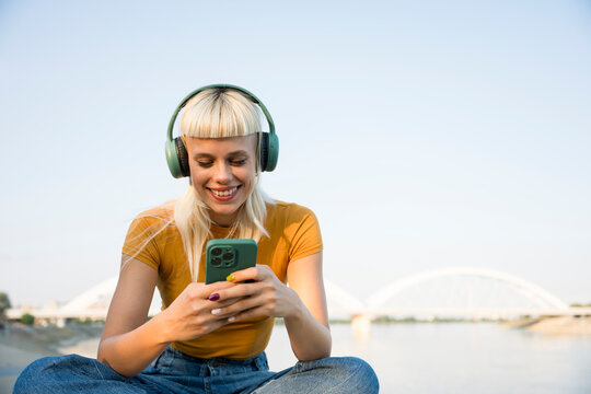Happy woman listening to music with headphones by riverside on phone