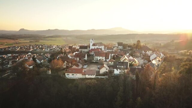 Lateral drone move to the left over Aach village at sunset, drifting past rooftops and the white church while warm golden light and distant hills create a serene panorama.