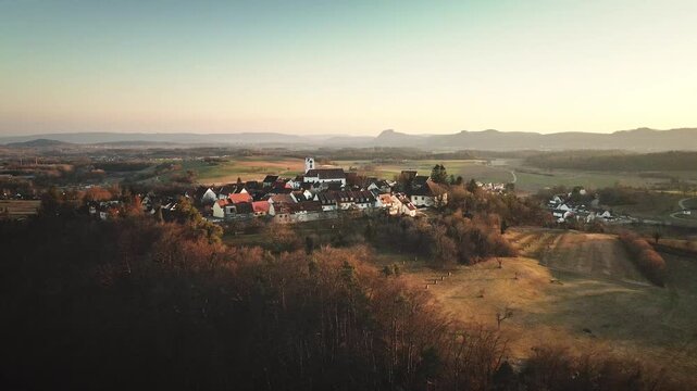 Forward-moving drone flight toward Aach village, approaching the white church as the surrounding fields and distant mountain ridge unfold in warm golden-hour light.