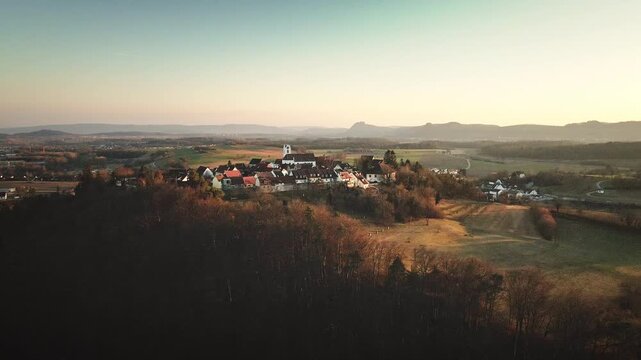 Forward-moving drone flight toward Aach village, approaching the white church as the surrounding fields and distant mountain ridge unfold in warm golden-hour light.
