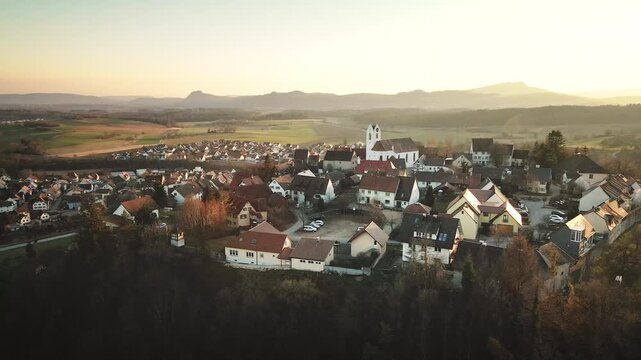 Orbiting drone shot circling Aach village around the white church and rooftops at golden hour, revealing the surrounding countryside and distant hills in warm sunset light.