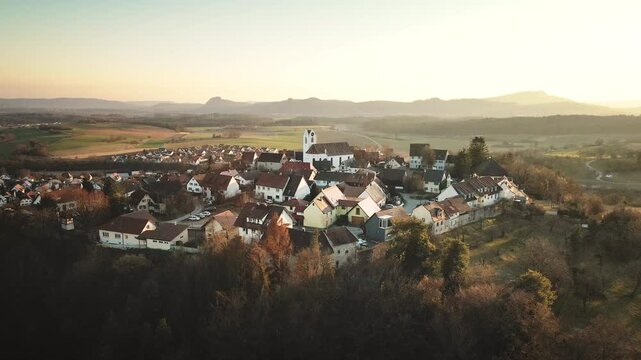 Lateral drone move to the left over Aach village at sunset, drifting past rooftops and the white church while warm golden light and distant hills create a serene panorama.