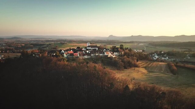 Forward-moving drone flight toward Aach village, approaching the white church as the surrounding fields and distant mountain ridge unfold in warm golden-hour light.