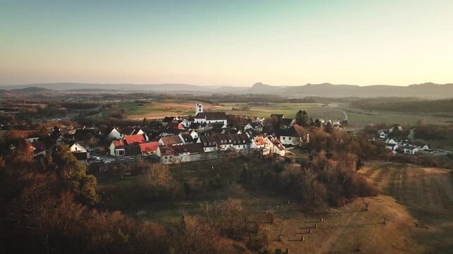 Drone flies backward away from Aach village, pulling back to reveal the hilltop rooftops, surrounding fields, and a wide mountain horizon bathed in warm golden-hour light.