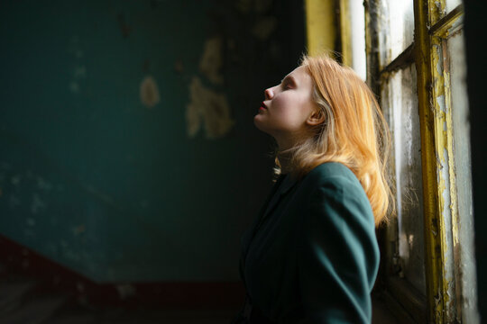 Red haired teenage girl in green jacket by old window with peeling paint