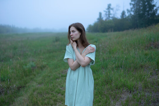 Woman in dress standing in foggy summer meadow near forest in morning