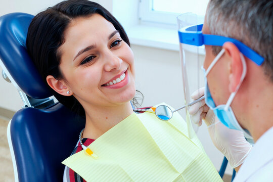Young adult Caucasian woman sitting in dental chair smiling while middle aged Caucasian male dentist wearing mask and face shield examining teeth with dental mirror and explorer