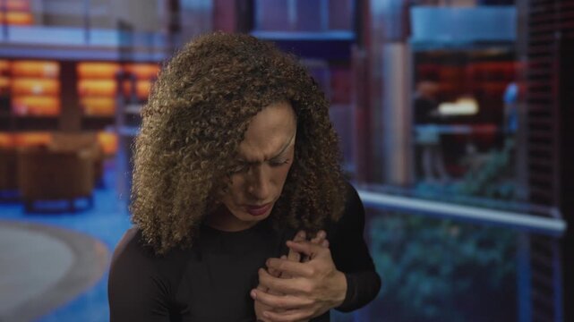 Transgender woman clutching chest in modern hotel lobby with tensed posture and pained expression; distress.