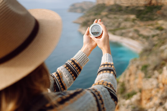 View from behind. Hiker in hat stands on edge of cliff, holding compass, overlooking sea. Young woman with compass on hiking trail enjoys scenery and feels sense of freedom. Navigation concept.