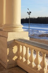Fototapeta premium 01.01.26. Moscow. Russia. Colonnade balcony overlooking snowy park path in Kuskovo. White stone pillars and balustrade with lamp post, dirt road and trees in winter background.