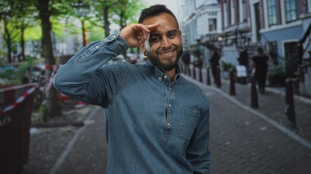 Man smiling with hand to forehead salute wearing denim shirt on a cobblestone city street with bollards and parked bicycles; confidence authenticity.