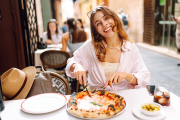 Portrait of a beautiful woman at a restaurant table, holding a slice of fragrant pizza. A cheerful woman enjoys a delicious Italian pizza at sunset. Concepts: food, tourism, enjoyment.
