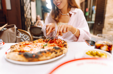 Portrait of a beautiful woman at a restaurant table, holding a slice of fragrant pizza. A cheerful woman enjoys a delicious Italian pizza at sunset. Concepts: food, tourism, enjoyment.